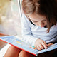 A young girl reading a picture book next to a window