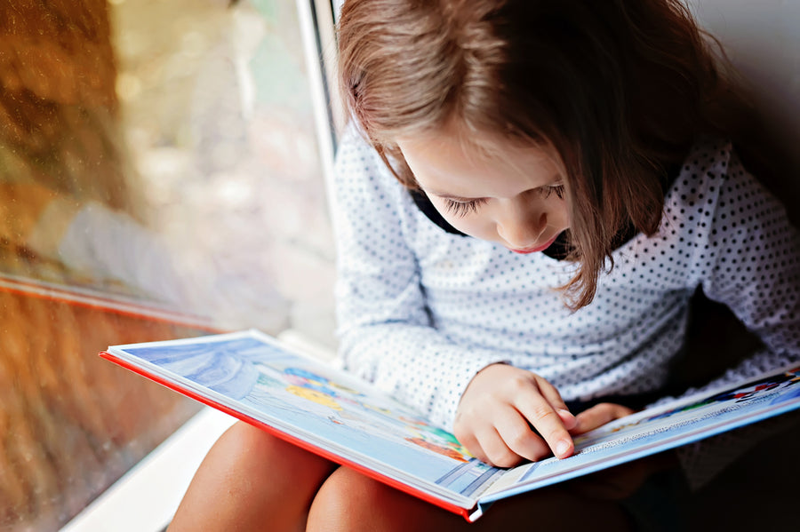 A young girl reading a picture book next to a window